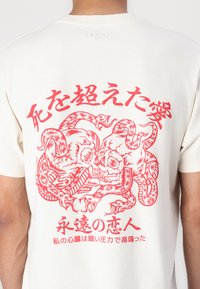 Biele bavlnené tričko s červeným grafickým dizajnom prepletených lebiek a hadov, s japonským textom tučným písmom na chrbte.