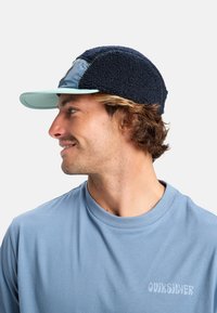 Cappello in pile blu navy con visiera azzurra e toppa. Presenta un tessuto testurizzato, dettagli di cucitura e un logo laterale in evidenza. T-shirt azzurra visibile.
