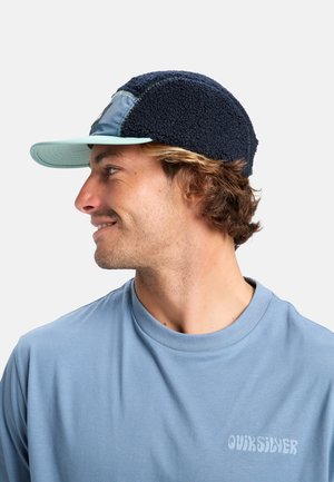 Casquette en polaire marine avec une visière bleu clair et un écusson. Présente un tissu texturé, des détails de couture et un logo sur le côté. T-shirt bleu clair visible.