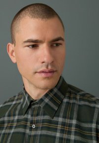 Chemise en tartan vert et gris avec un col boutonné. Le tissu semble doux avec une légère texture, comportant des lignes jaunes en accent dans le motif.