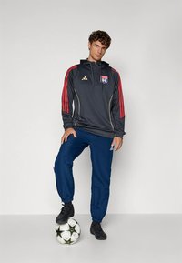 Hoodie de sport gris avec des rayures rouges et dorées, arborant un logo, associé à un pantalon bleu et des chaussures de football noires, debout sur un ballon.