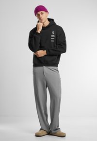 Zwarte hoodie met logo's, grijs op maat gemaakte broek en beige slippers. De hoodie heeft een voorzak en een trekkoordcapuchon. Roze beanie op het hoofd.