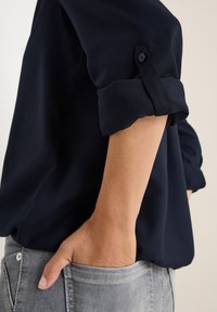 Marinebluse med oprullede ærmer, glat tekstur og knapdetalje, parret med lysegrå jeans med møntlomme.