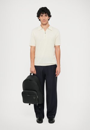Mochila de cuero negro con una textura suave, bolsillo frontal con cremallera y correas ajustables, combinada con una camiseta tipo polo crema y pantalones a rayas.