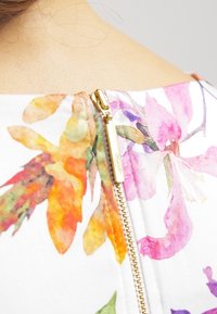 Tissu à imprimé floral aux couleurs vives, accompagné d'une fermeture éclair dorée avec une étiquette logo, mettant en avant une texture lisse et une finition douce.