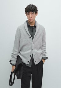 Ljusgrå rillad cardigan med sjalkrage, svarta knappar och en strukturerad finish, i lager över en mörkgrå skjorta. Svarta byxor och väska.