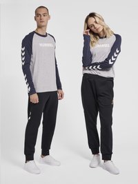 Herrgrå långärmad skjorta med marinblå ärmar och "HUMMEL"-logotyp; svarta joggers med sidofickor; båda bär vita sneakers.