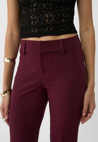 Burgundowe dopasowane spodnie o gładkiej fakturze, z płaskim przodem i dwoma bocznymi kieszeniami. Połączone z czarnym koronkowym topem na krótki rękaw i złotymi akcesoriami.