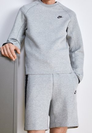 Mann trägt graues Nike-Sweatshirt und passende Shorts, steht mit einer Hand auf einem grauen Schrank vor weißem Hintergrund.