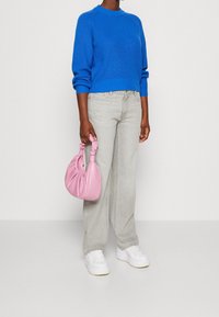 Pull tricot bleu vif, jean gris clair, baskets blanches, tenant un sac à main rose texturé avec des accents froncés. Fond neutre.