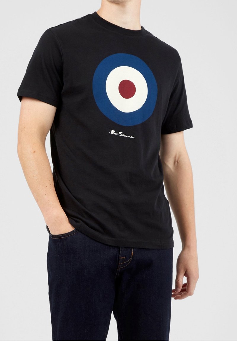 Camiseta de algodón negra con un gran gráfico circular en azul, blanco y rojo. Con un estilo de mangas cortas y un corte casual. Nombre de la marca mostrado abajo.