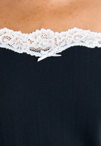 Maglia nera lavorata a maglia con un bordo in pizzo bianco sul collo, decorata con un piccolo fiocco al centro.
