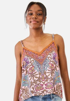 Joven mujer con una blusa sin mangas de tirantes finos y estampado floral colorido, y vaqueros azules, de pie frente a un fondo blanco.