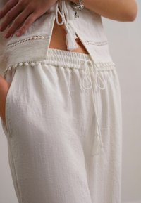 Ensemble en lin blanc comprenant un haut léger avec des détails en dentelle et un pantalon à cordon avec des pampilles et une taille froncée.