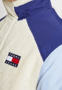 Pullover in pile con sezioni blu navy, azzurro e crema; presenta una toppa con il logo "TOMMY JEANS" e bottoni a pressione al colletto.