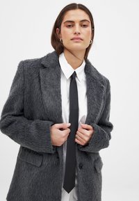 Flauschiger grauer Blazer mit Reverskragen, zwei vorderen Taschen und einem Einzelknopfverschluss, über einem weißen Hemd und einer schwarzen Krawatte getragen.