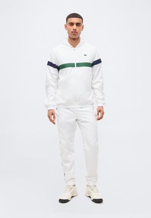 Férfi áll fehér melegítőben, amelyen zöld és tengerészkék csíkok futnak a mellkason és a karokon, fehér edzőcipőben, sima háttér előtt.