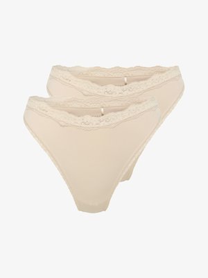 Kobieta w beżowym biustonoszu z koronkową lamówką oraz dopasowanych, wysokich figach, stojąca na tle gładkiego, jasnego tła.