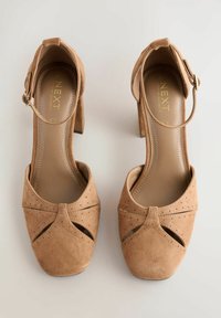 Beige Wildleder-Pumps mit geschlossenem Zehenbereich, geschwungenen Aussparungen und Knöchelriemen. Mit Perforationsdetails und einer strukturierten Oberfläche gestaltet.
