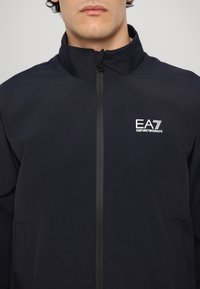 Chaqueta ligera de color azul marino con cuello alto, cremallera frontal y detalle del logo en el pecho. Textura suave, diseño minimalista, sin patrón.