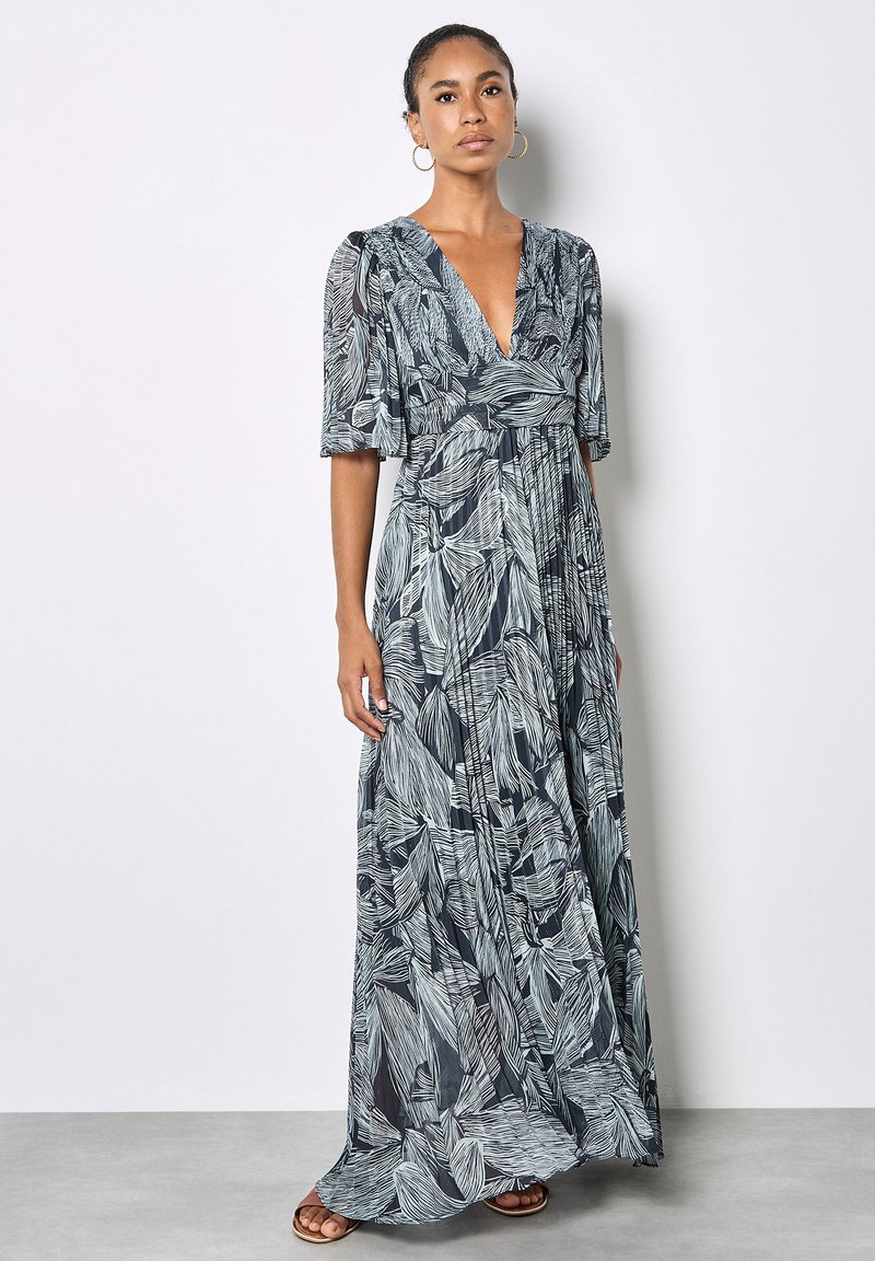 Femme debout portant une robe longue noire avec des motifs de feuilles blanches, un décolleté en V, des manches mi-longues et des boucles d'oreilles créoles ainsi que des sandales.