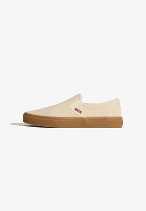 Zapatilla beige tipo slip-on de Vans con suela de goma marrón y pequeña etiqueta roja con el logo de Vans cerca del panel lateral elástico.