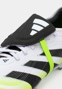 Cizma de fotbal albă și neagră, cu un upper texturat, având o limbă neagră lucioasă și o curea de accent verde neon. Logo-ul cu trei dungi este vizibil.