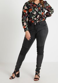 Femme portant un chemisier noir à fleurs à manches longues, un jean skinny côtelé gris foncé et des sandales à talons blocs ouvertes noires, posant avec une main sur la hanche.