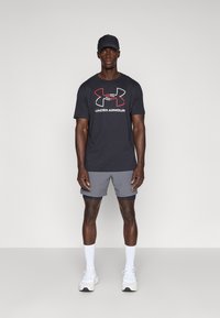 Svart kortärmad T-shirt med röd och vit Under Armour-logotyp, tillsammans med grå shorts och vita sportstrumpor. Modellen bär en svart keps.