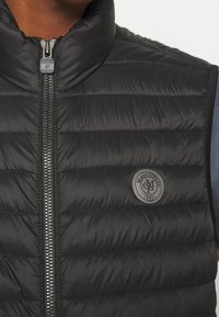 Schwarze gesteppte Weste mit hohem Kragen, Vorderreißverschluss und einem runden Logo-Patch. Verfügt über horizontale Steppnähte und eine glatte Textur.