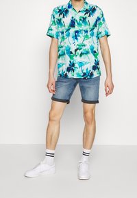 Koszulka z krótkim rękawem w jasnym niebieskim kolorze z zielonym i czarnym abstrakcyjnym wzorem kwiatowym, w połączeniu z dżinsowymi szortami i białymi trampkami w paski.