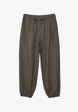 Mladá žena s kučeravými vlasmi, ktorá má na sebe tmavohnedý bezrukávový halter top a voľné nohavice, kráča vpred s jednou rukou v vrecku.