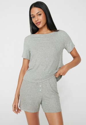 Frau mit langen schwarzen Haaren, die ein hellgraues kurzärmeliges Oberteil und passende Shorts trägt, steht vor einem einfarbigen weißen Hintergrund.