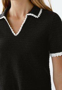 Haut noir texturé avec un col en V orné d'un bord festonné blanc au niveau du col et des manches courtes, souligné par un bord similaire aux manches.