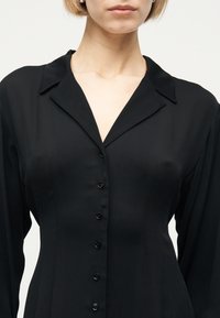 Γυναίκα που φοράει εφαρμοστή μαύρη μπλούζα με μακριά μανίκια, γιακά λάπι, και επτά κουμπιά μπροστά, εμφανιζόμενη από τον λαιμό έως τη μέση.