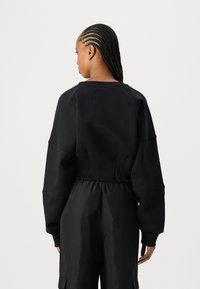 Svart sweatshirt med avslappet passform, vide ermer og ribbet halslinning. Glatt stoff og subtile sømmer detaljerer bakdesignet. Kombinert med svarte bukser.