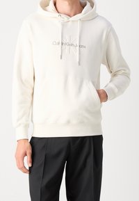 Vit bomullshoodie med ficka fram, dragskor och broderad logo "CK" i mitten. Ribbad mudd och fåll, kombinerad med svarta byxor.
