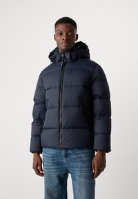 Navy puffer jacket with a hood, zip closure, and elastic cuffs. Textured, quilted design with side pockets. Worn over blue denim jeans.