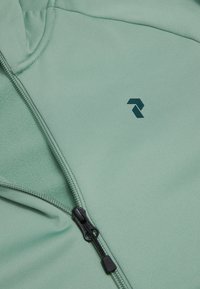 Lichtgroene jas met ritssluiting, zwarte rits en klein zwart logo op de borst, met zichtbare gladde en fleece stoftexturen.