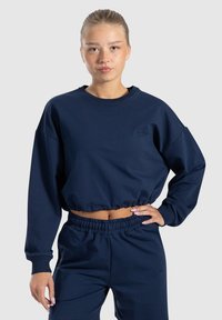 Felpa cropped blu navy con maniche lunghe e polsini a costine, caratterizzata da un logo ricamato. Abbinata a pantaloni con vita elasticizzata dello stesso colore.