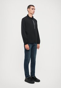 Casaco preto com fecho zip, com gola, apresentando um bolso frontal, usado sobre jeans azul escuro e ténis pretos. Tecido suave com design aerodinâmico.