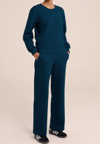 Tealfärgad sweatshirt och matchande byxor av strukturerad tyg. Sweatshirten har rund halsringning och ribbade ärmmuddar; byxorna har sidofickor.