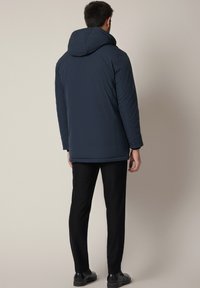 Mężczyzna ubrany w ciemnogranatową kurtkę z kapturem, czarne spodnie i czarne buty, stojący w kierunku jasnobeżowej ściany.