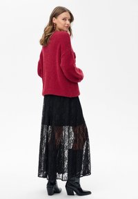Czerwony sweter z szerokimi rękawami, połączony z czarną koronkową maxi spódnicą z przezroczystymi panelami oraz czarnymi botkami do kostek.
