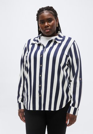 Frau mit geflochtenem Haar, die ein schwarz-weiß gestreiftes Hemd mit Knöpfen und schwarze Hosen vor einem einfachen Hintergrund trägt.