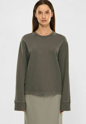 Kvinde med glat brunt hår iført løs mørkegrå langærmet sweatshirt og lys beige nederdel, stående foran en ensfarvet baggrund.