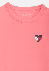 Ροζ παιδικό πουκάμισο με κουμπί ασφαλ είας στον ώμο, λογότυπο Tommy Hilfiger μέσα στον γιακά και μια μικρή δίχρωμη καρδιά με χαμογελαστά προσωπάκια στο στήθος.