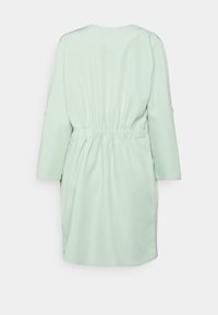 Pistacjowa sukienka o luźnym kroju, z podkreśloną talią, rękawami o długości trzy czwarte i gładką fakturą. Brak widocznych wzorów lub ozdób.