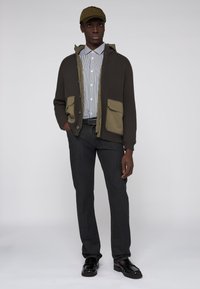 Grön ribbade cardigan med två stora insticksfickor över en randig skjorta med knappar, i kombination med mörka jeans och svarta loafers, avslutat med en keps.