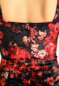 Rückansicht einer Person, die ein schwarzes Samtkleid mit rotem und beigem Blumenmuster und einem zentralen schwarzen Reißverschluss trägt.